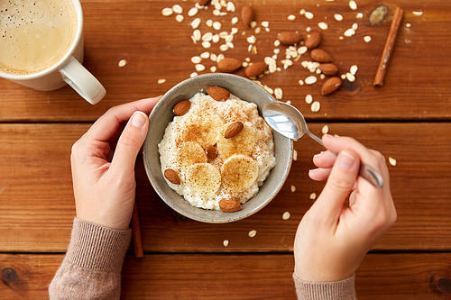 food and breakfast concept - hands of woman eating oatmeal porridge in bowl with sliced ​​banana, almond nuts and cinnamon and cup of coffee on wooden table