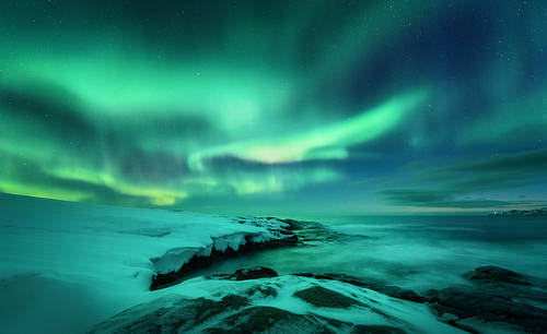 Aurora borealis over ocean. Northern lights in Teriberka, Russia. Starry sky with polar lights and clouds. Night winter landscape with aurora, sea with stones in blurred water, snowy mountains. Travel