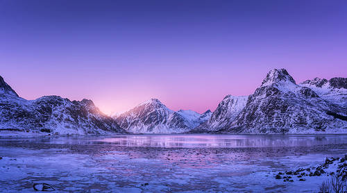 Snowy mountains, blue sea with frosty coast, reflection in water and purple sky at colorful sunset in Lofoten islands, Norway. Winter landscape with snow covered rocks, fjord with ice at night. Nature