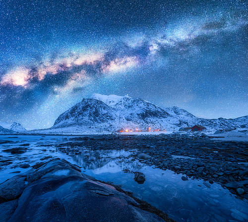 Milky Way over the snow covered mountains and rocky beach in winter at night. Lofoten Islands, Norway. Landscape with blue starry sky, water, stones, snowy rocks, bright galaxy and city lights. Space