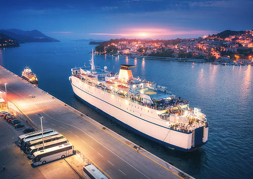 Aerial view of cruise ship in port at sunset. Landscape with ships and boats in harbor, city lights, buildings, mountains, blue sea at night. Top view. Luxury cruise. Floating liner at harbor at dusk