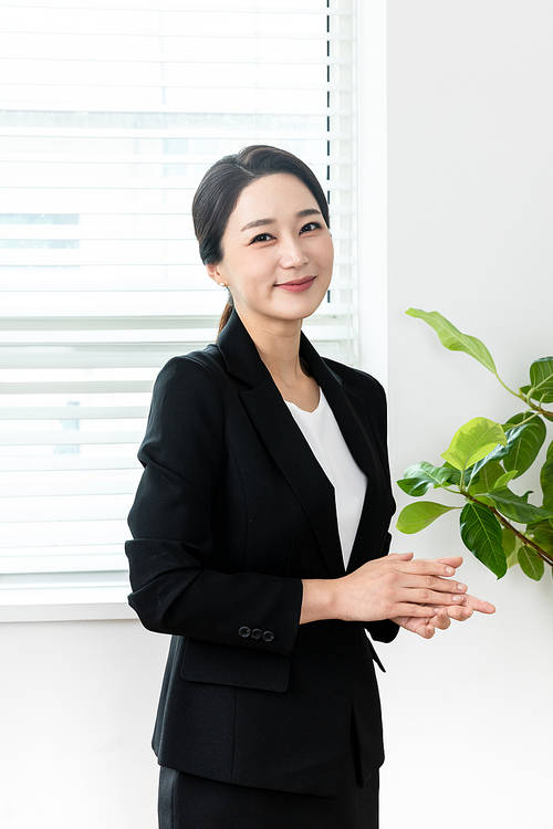 A scene image of a woman in a black suit looking straight ahead with a smile