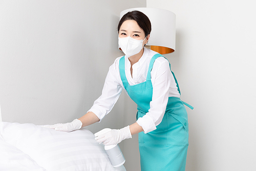 A scene image of a woman wearing an apron wearing a mask and disinfecting the bed