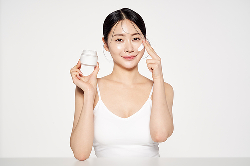 Asian woman holding a cream container and applying cosmetics to her face