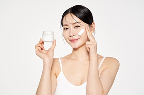 Asian woman holding a cream container and applying cosmetics to her face