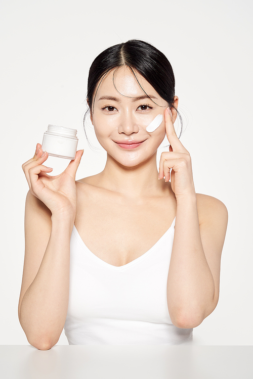 Asian woman holding a cream container and applying cosmetics to her face