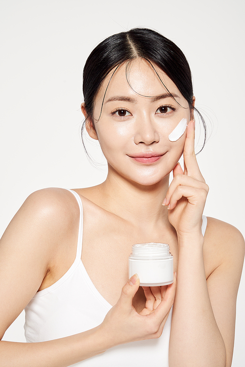 Asian woman holding a cream container and applying cosmetics to her face