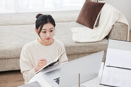 A woman opens her notebook, looks at her laptop, and takes notes with a pen