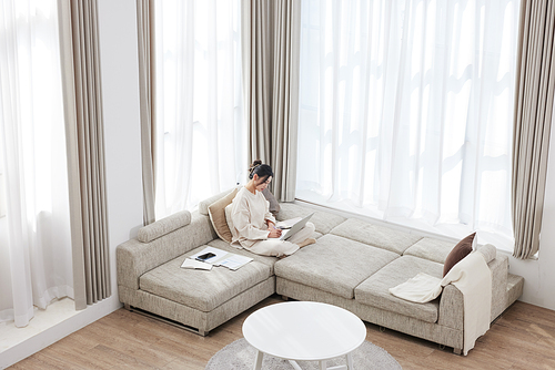 A woman sitting cross-legged on the sofa, taking notes and working on a laptop