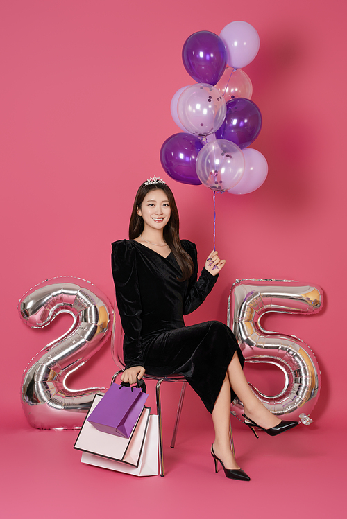 A woman in her 20s sitting and posing with a balloon and a shopping bag in front of a 25 number balloon.