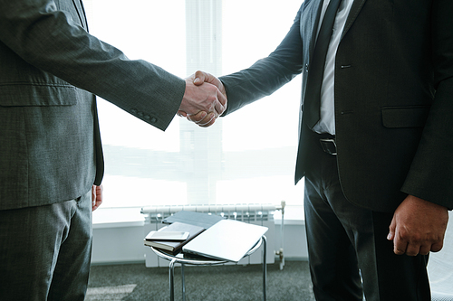 Midsection of two successful business partners in formalwear standing against small table with notebook and gadgets during handshake