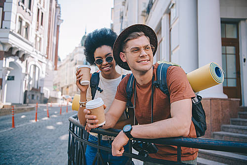 smiling multicultural couple of tourists standing with disposable cups of coffee