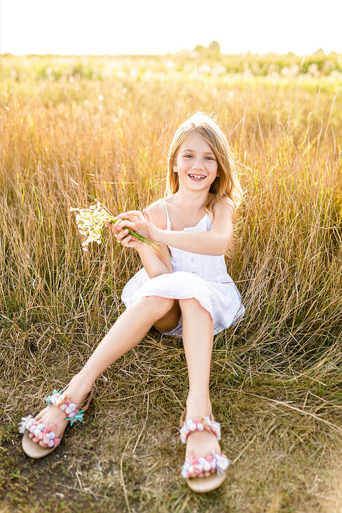 happy little child in white dress with field flowers bouquet sitting in field