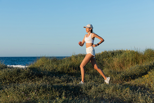 sportive woman in and wireless earphone running on grass near sea