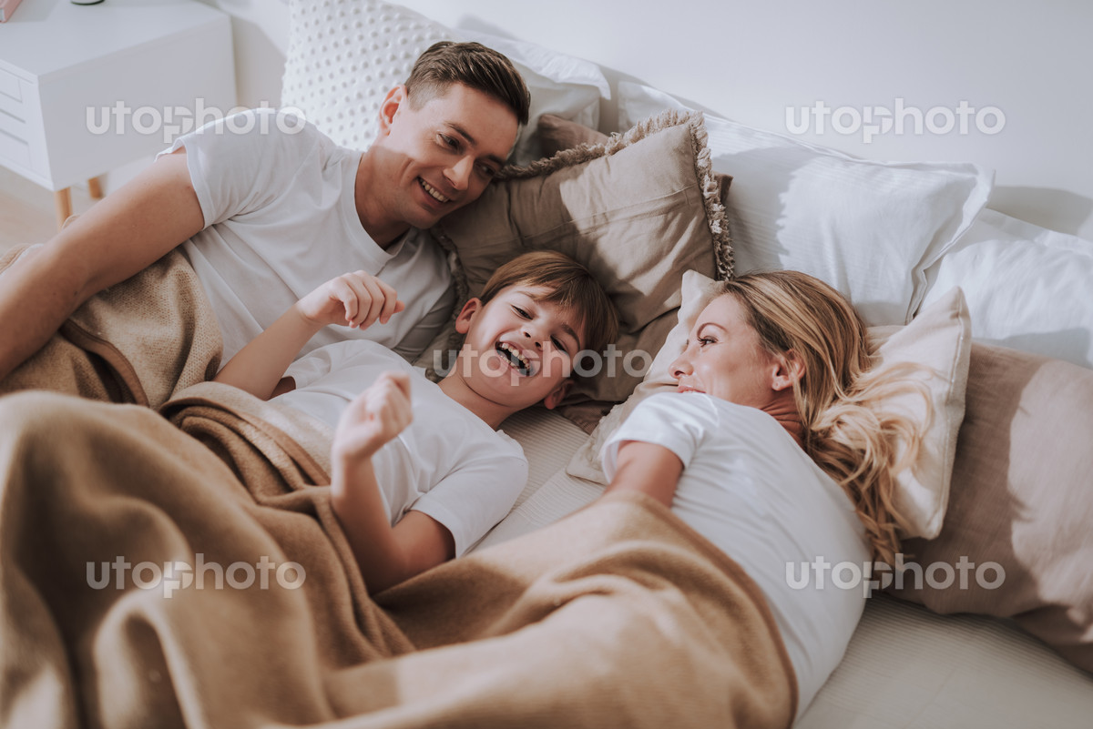 Positive morning mood. Emotional kid laughing and feeling happy while lying in comfortable bed between his parents